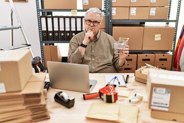 Sticker - Senior caucasian man working at small business ecommerce holding shopping cart serious face thinking about question with hand on chin, thoughtful about confusing idea