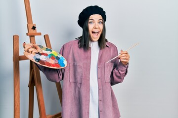 Wall Mural - Young brunette woman standing by empty easel stand holding palette celebrating crazy and amazed for success with open eyes screaming excited.