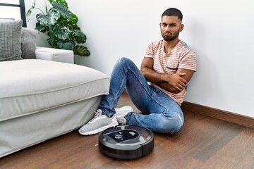 Wall Mural - Young indian man sitting at home by vacuum robot skeptic and nervous, disapproving expression on face with crossed arms. negative person.