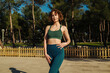 Beautiful young redhead woman wearing green sports bra and blue yoga pants poses to camera. Sportive woman looking at the camera while standing on city park, outdoors.
