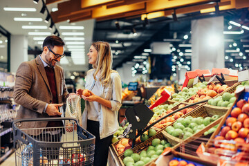 Wall Mural - Loving couple in a supermarket with a shopping trolley and mesh bag choosing fruits