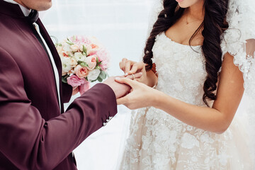The bride wears a wedding ring to the groom at the wedding ceremony. The groom in a crimson suit with a boutonniere, with a bouquet in his hands, the bride is dressed in a light dress with patterns.