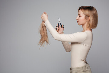 Wall Mural - Studio portrait of blonde female cutting her own hair, hairstyle and haircut, lock of hair, scissors in hand, plain background, isolated, changes and hair treatment concept, lifestyle beauty and style