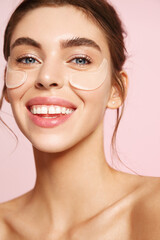 Wall Mural - Close up of beauty girl with natural no makeup look and white smile, has silicone under eye patches, smiling happy, using skincare products from dark circles and puffiness, pink background