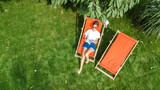Fototapeta Sawanna - Young girl relaxes in summer garden in sunbed deckchair on grass, woman reading book outdoors in green park on weekend, aerial drone view from above
