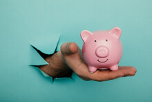 Hand With A Pink Piggy Bank Through A Blue Paper Hole. Financial And Business Concept