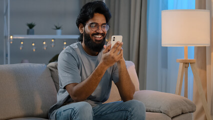 Happy arabian indian male millennial man holding modern smartphone texting message at home sitting on couch in evening bearded guy in glasses using mobile app watching funny video online laughing