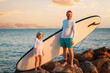 © _KUBE_ - Summer activity vacations. Happy father and daughter standing with sup board on big beach rocks. Sunset sky and ocean at the background. Surfing