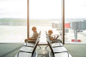 Children play games on tablet or smartphone, while waiting for their airplane flight in transit hall near departure gate in empty airport. Family travel by air with child.