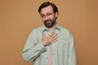 Studio portrait of bearded man standing over beige background wears casual shirt and glasses keeps his palm on heart and smiles