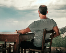 An Elderly Atletic Male Sitting At Old Wooden Table And Looking At Cloudy Sky