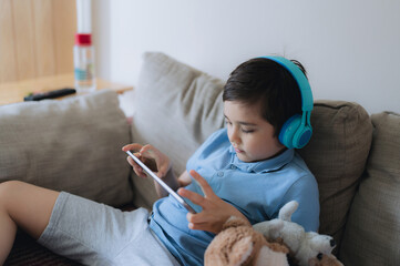 Wall Mural - Happy young boy wearing headphone for playing game on internet with friends, Child sitting on sofa reading or watching cartoon on tablet, School kid doing homework online at home.