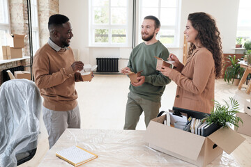 Group of positive young multi-ethnic employees standing in circle and chatting during lunch break in new office with furniture under polyethylene