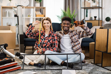 Wall Mural - Happy multicultural couple sitting on floor at living room, looking at camera and holding various tools for assembling furniture. Concept of young family moving in new house.