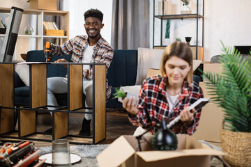 Wall Mural - African american man using hand drill for assembling wooden shelves while his caucasian wife unpacking boxes after moving to new apartment. Family and new chapter in life concept.