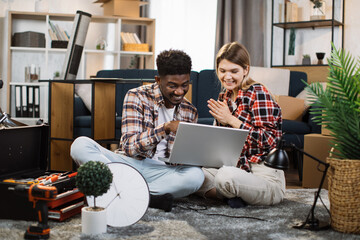 Wall Mural - Cheerful young man and woman in casual attire smiling sincerely while looking on laptop screen. Beautiful family sitting on floor at living room with cardboard boxes around.