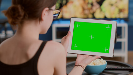 Wall Mural - Closeup of young woman holding digital tablet with green screen watching webinar sitting on sofa in front of tv. Girl looking at touchscreen device with chroma key attending online course at home.