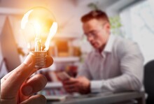 Human Hand Holds Light Bulb On Workplace Background. Save Energy Concept.