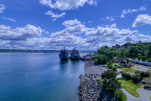 Two Ships Docked On The Rocky Shores Of Puget Sound In Tacoma, Washington Under A Blue Cloud Filled Sky.