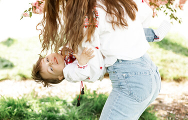 Little boy on hands of mother. Woman playing with child outside in blooming spring garden. Portrait of family of two people. Spring landscape background.