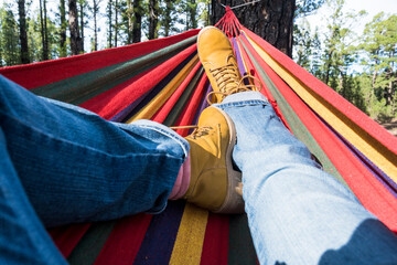 Close up of pair of trekker legs having relax outdoor leisure activity laying on a colorful hammock alone with tress forest woods in background and blue sky