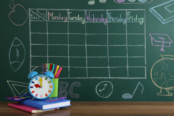 Canvas Print - Alarm clock and stationery on wooden table near green chalkboard with drawn school timetable