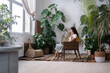 © DimaBerlin - Smiling young female owner city greenhouse sitting on chair in front of window with laptop having rest, buying plants online, looking intresting serias, texting with friends. Monstera on background.