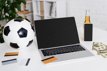 laptop with blank screen near soccer ball, credit cards, dollars and bottle of beer.