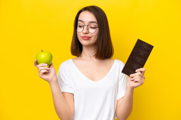 Young Ukrainian woman isolated on yellow background having doubts while taking a chocolate tablet in one hand and an apple in the other