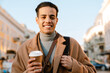Young man smiling and drinking coffee while walking at city street