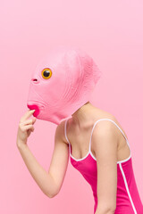 Wall Mural - A funny woman wearing a pink fish mask on her head put her fingers in her mouth with pink clothes on a pink background. The concept of modern art photography