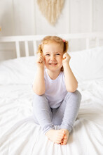 A Little Cute Girl Child On A White Cotton Bed At Home Is Fooling Around Playing With Tails And Smiling, The Child Is Playing And Messing Around At Home In The Morning
