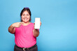 Young curvy latina woman smiling holding and pointing at her mobile phone, isolated over blue background. Copy space