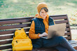 Child caucasian teen boy in yellow beany hat and backpack sitting on a bench outdoors using laptop. Remote or distant learning on the go. Blogging surfing. Homeschooling. Video call. Copy space.