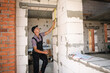 © Ольга Симонова - Construction worker at construction site measures the length of window opening and brick wall with tape measure. Cottage are made of porous concrete blocks, work clothes - jumpsuit and baseball cap