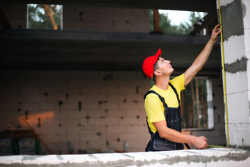 Construction worker at construction site measures the length of window opening and brick wall with tape measure. Cottage are made of porous concrete blocks, work clothes - jumpsuit and baseball cap