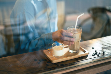 Wall Mural - A hand holding a tall glass of iced latte coffee with milk on a wooden bar over a cafe glass window reflex at a Cafe coffee shop. Cold brew refreshment summer drink with copy space. Selective focus