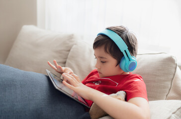 Wall Mural - Happy young boy wearing headphone for playing game on internet with friends, Child sitting on sofa reading or watching cartoon on tablet, School kid doing homework online at home.