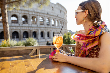 Woman drinking Spritz Aperol at outdoor cafe near Coliseum, the most famous landmark in Rome. Concept of italian lifestyle and traveling Italy. Caucasian woman wearing dress and shawl in hair