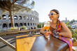 Woman drinking Spritz Aperol at outdoor cafe near Coliseum, the most famous landmark in Rome. Concept of italian lifestyle and traveling Italy. Caucasian woman wearing dress and shawl in hair