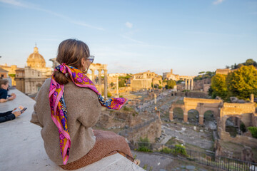 Wall Mural - Woman enjoying view on the Roman Forum, ruins at the center of Rome on a sunset. Concept of traveling famous landmarks in Italy. Caucasian woman wearing colorful shawl in hair and sunglasses