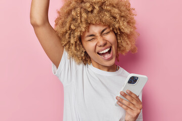 Wall Mural - Excited cheerful young woman makes winner gesture keeps arm raised up holds mobile phone gets excellent news dressed in casual white t shirt isolated over pink background. Celebration concept