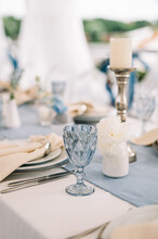 Long Dinner Tables Covered With White Cloth, Served With Porcelain And Blue Glasses And Rich Decorated With Flowers 
