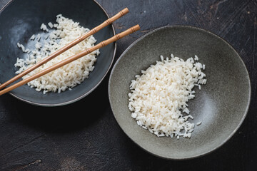 Two plates with steamed white rice and bamboo chopsticks on a dark-brown stone background, horizontal shot, elevated view