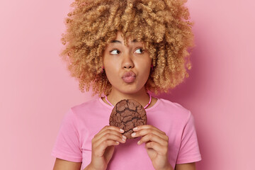 Wall Mural - Portrait of good looking curly haired young woman keeps lips folded holds delcious chocolate cookie wants to eat sweet food dressed in casual t shirt isolated over pink background. Snack time