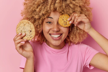 Wall Mural - Positive carefree curly haired woman has sweet tooth holds delicious cookies over eye smiles pleasantly dressed in casual t shirt glad to eat delicious snack isolated over pink studio background.