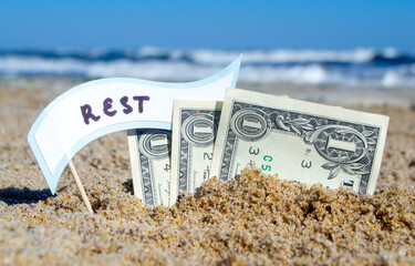 Paper bills one dollars, small stick with paper speech bubble with word Rest buried in sand beach background sea close-up in sunny summer day. Concept Money travel tourism vacation holiday relax