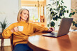 © baranq - Mid adult woman using laptop at home sitting at a table with cup of coffee
