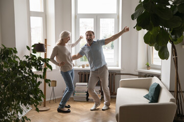 Excited happy senior couple dancing to music together at home couch, enjoying date, anniversary, active entertainment, party for two, having fun together. Retirement, indoor activity concept