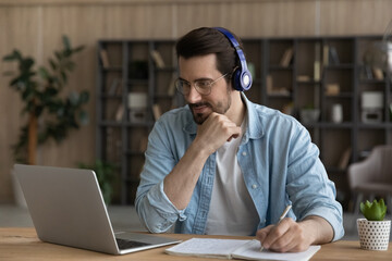 Remote studying. Concentrated millennial male in modern earphones watch video course on laptop take notes learn virtual lesson. Busy young guy take part at online training write up useful information